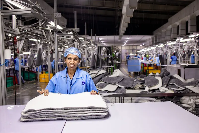 Trabalhadora sorrindo em uma fábrica clara, com colegas ao fundo